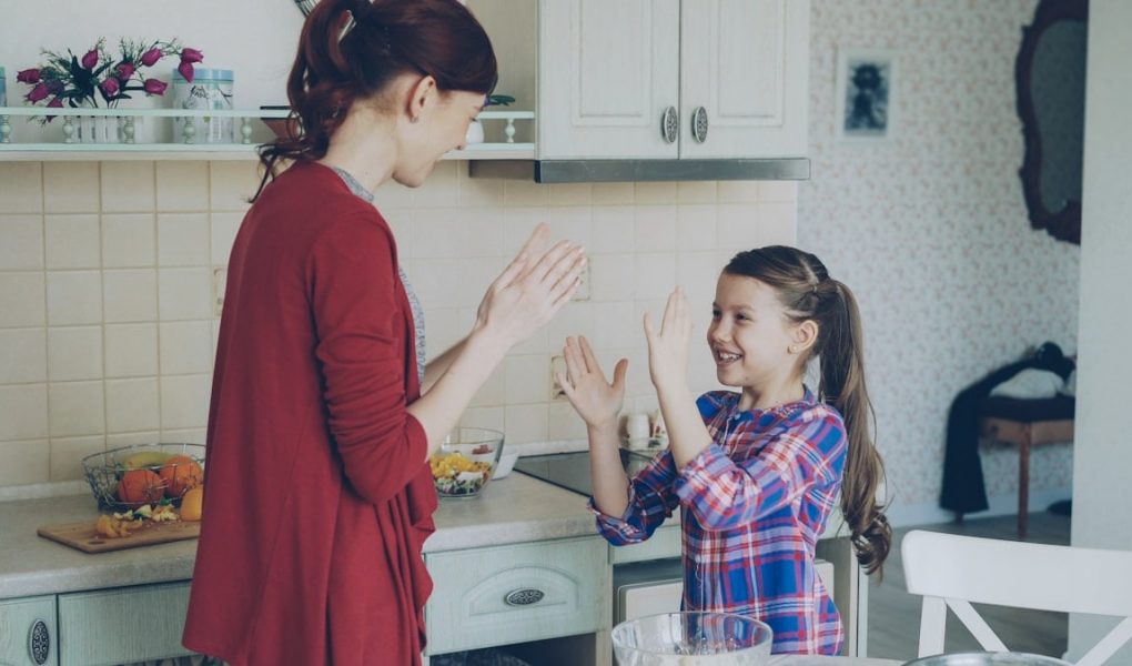 Happy young mother and cute funny daughter playing clapping hands game and have fun while cooking in kitchen at home. Family, cooking and people concept