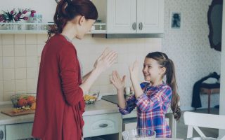 Happy young mother and cute funny daughter playing clapping hands game and have fun while cooking in kitchen at home. Family, cooking and people concept