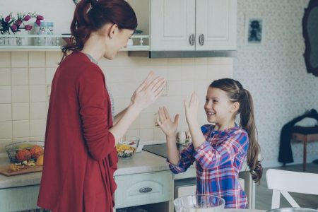 Happy young mother and cute funny daughter playing clapping hands game and have fun while cooking in kitchen at home. Family, cooking and people concept